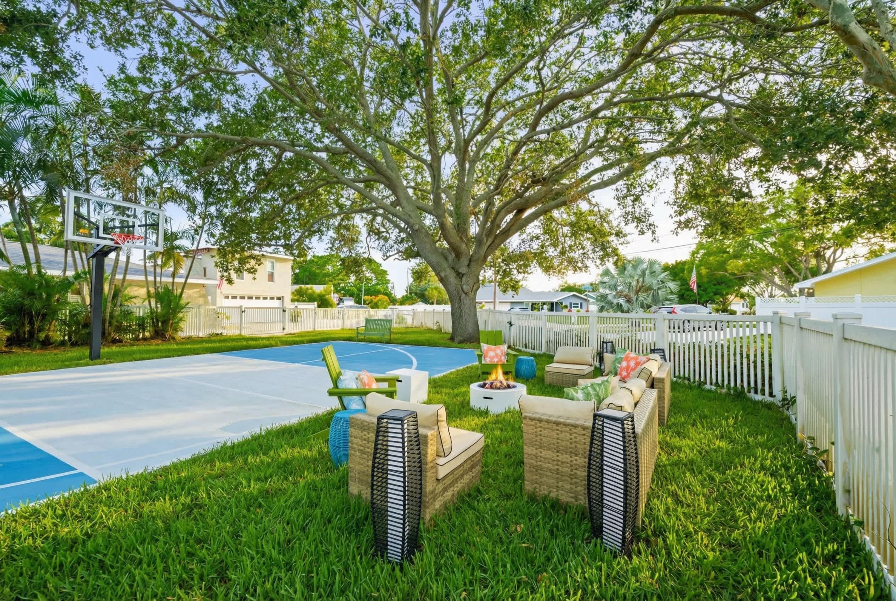 A backyard with a basketball court, outdoor seating, and a large tree.