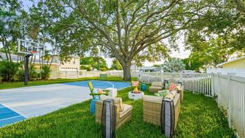 A backyard with a basketball court, outdoor seating, and a large tree.