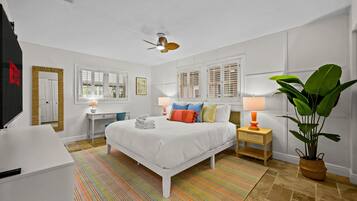 A hotel room with a bed, bedside tables, a desk, a chair, a ceiling fan, windows with shutters, a large plant, and a striped rug.
