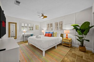 Habitación de hotel con cama, mesitas de noche, escritorio, silla, ventilador de techo, ventanas con contraventanas, una planta grande y una alfombra a rayas.