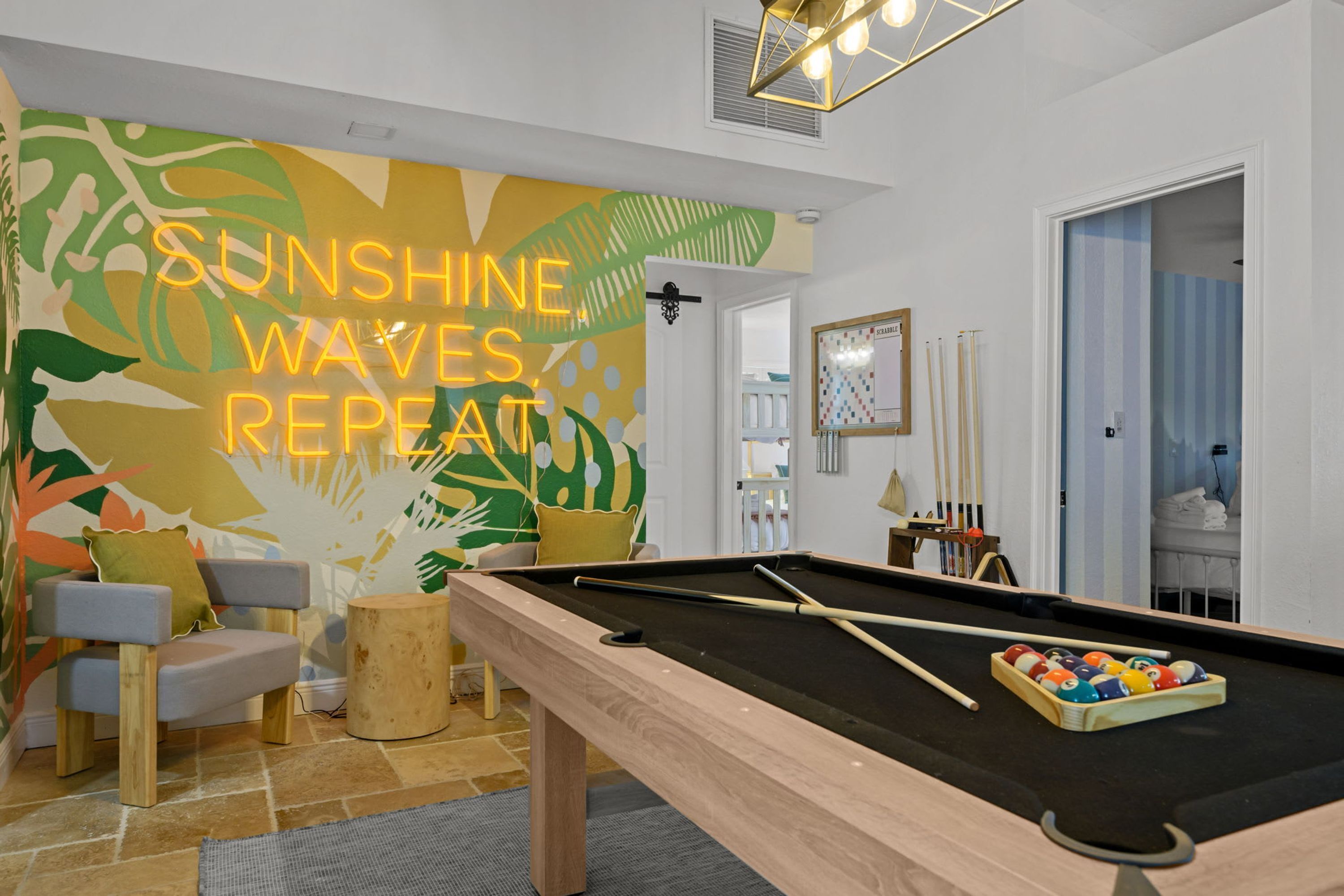 A pool table with a pool cue and balls, a sofa, a small wooden stool, and a wall with a colorful abstract design.