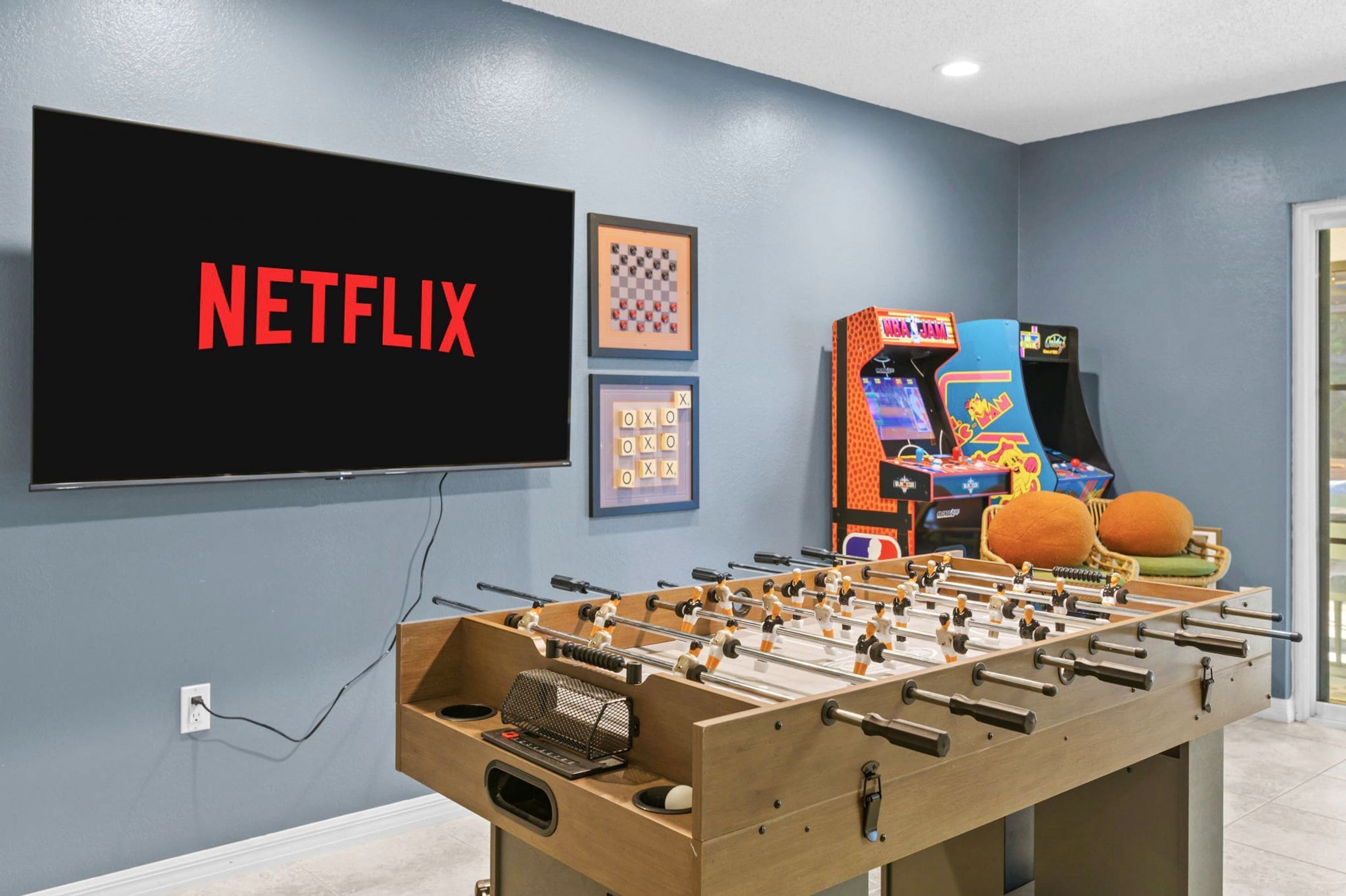 Una habitación con mesa de futbolín, televisor mostrando 'NETFLIX' y juegos de arcade.