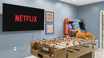 Una habitación con mesa de futbolín, televisor mostrando 'NETFLIX' y juegos de arcade.