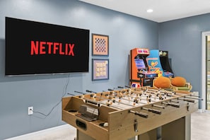 Una habitación con mesa de futbolín, televisor mostrando 'NETFLIX' y juegos de arcade.