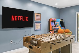 Una habitación con mesa de futbolín, televisor mostrando 'NETFLIX' y juegos de arcade.