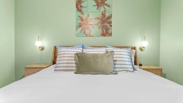 Una cama con sábana blanca, dos mesitas de noche, dos lámparas de pared y una obra de arte con motivos tropicales.
