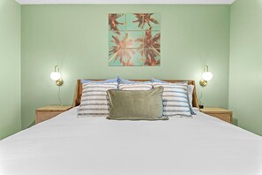 Una cama con sábana blanca, dos mesitas de noche, dos lámparas de pared y una obra de arte con motivos tropicales.