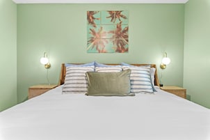 Una cama con sábana blanca, dos mesitas de noche, dos lámparas de pared y una obra de arte con motivos tropicales.