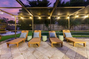 Un área de descanso al aire libre con tumbonas y cojines de madera, bajo una estructura cubierta con luces colgantes.