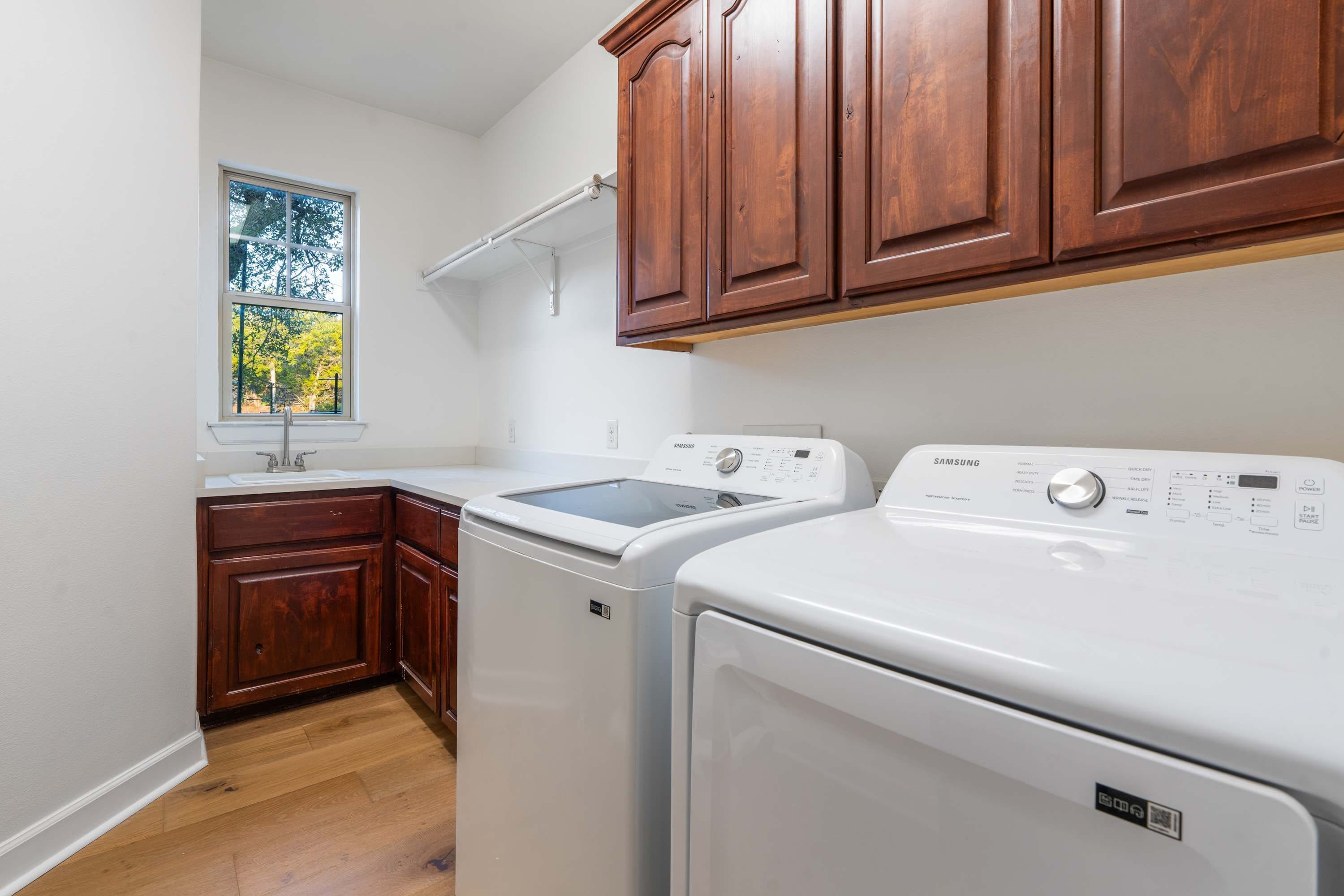 A laundry room with two white washers and dryers, wooden cabinets, a window, and a white sink.