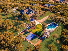 An aerial view of a residential property with a swimming pool, tennis court, and a large lawn area.