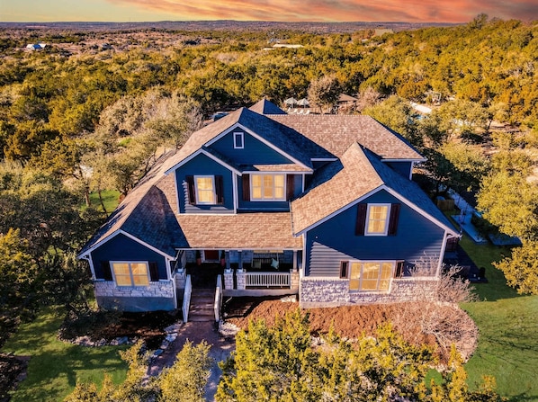 A two-story house with a front porch and a garage, surrounded by trees and a hilly landscape. - Dink Dip Dream - Pickleball - Pool - Putt Putt (Austin)