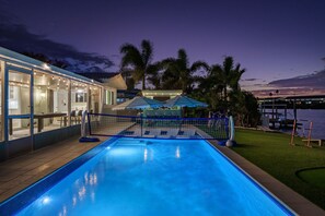 A swimming pool with a netted area, surrounded by a building with glass doors and windows, palm trees, and a view of the water. - Waterfront Walk to Beach Pool Kayaks Bikes (Indian Rocks Beach)