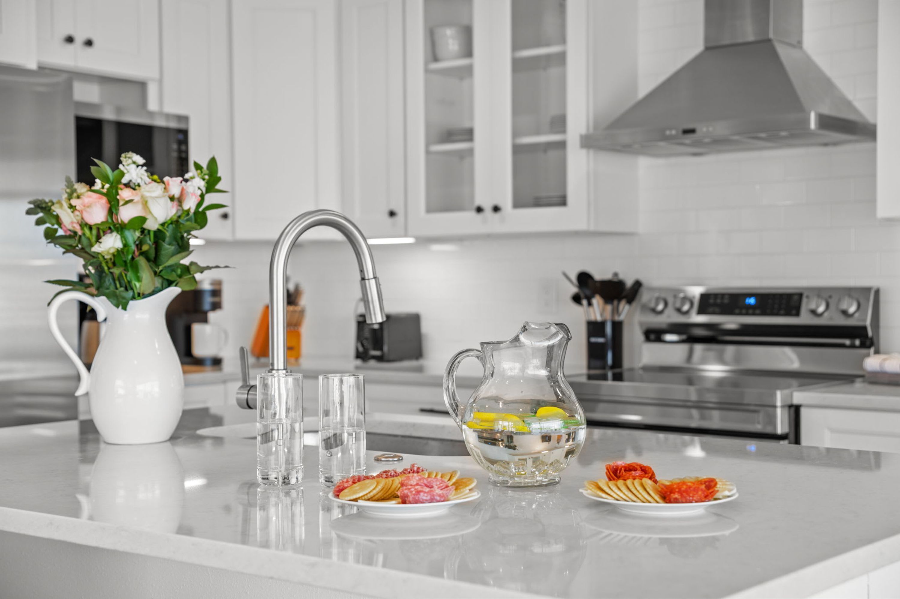 A modern kitchen with white cabinetry, a stainless steel range hood, and a central island with a vase of flowers, glasses, and plates of food.