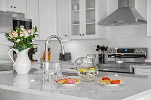 A modern kitchen with white cabinetry, a stainless steel range hood, and a central island with a vase of flowers, glasses, and plates of food. - Waterfront Walk to Beach Pool Kayaks Bikes (Indian Rocks Beach)