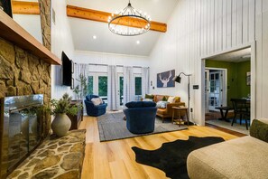 A spacious living room with a stone fireplace, a chandelier, and a view of the outdoors through large windows. - Modern Peaks Hot Tub Sauna Theatre Room (McGaheysville)