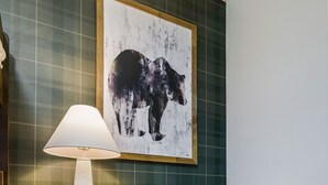 Una habitación con un papel tapiz estampado en cuadros, un cuadro abstracto enmarcado, una mesita de noche de madera con una lámpara y un sillón de cuero con una manta de piel.
