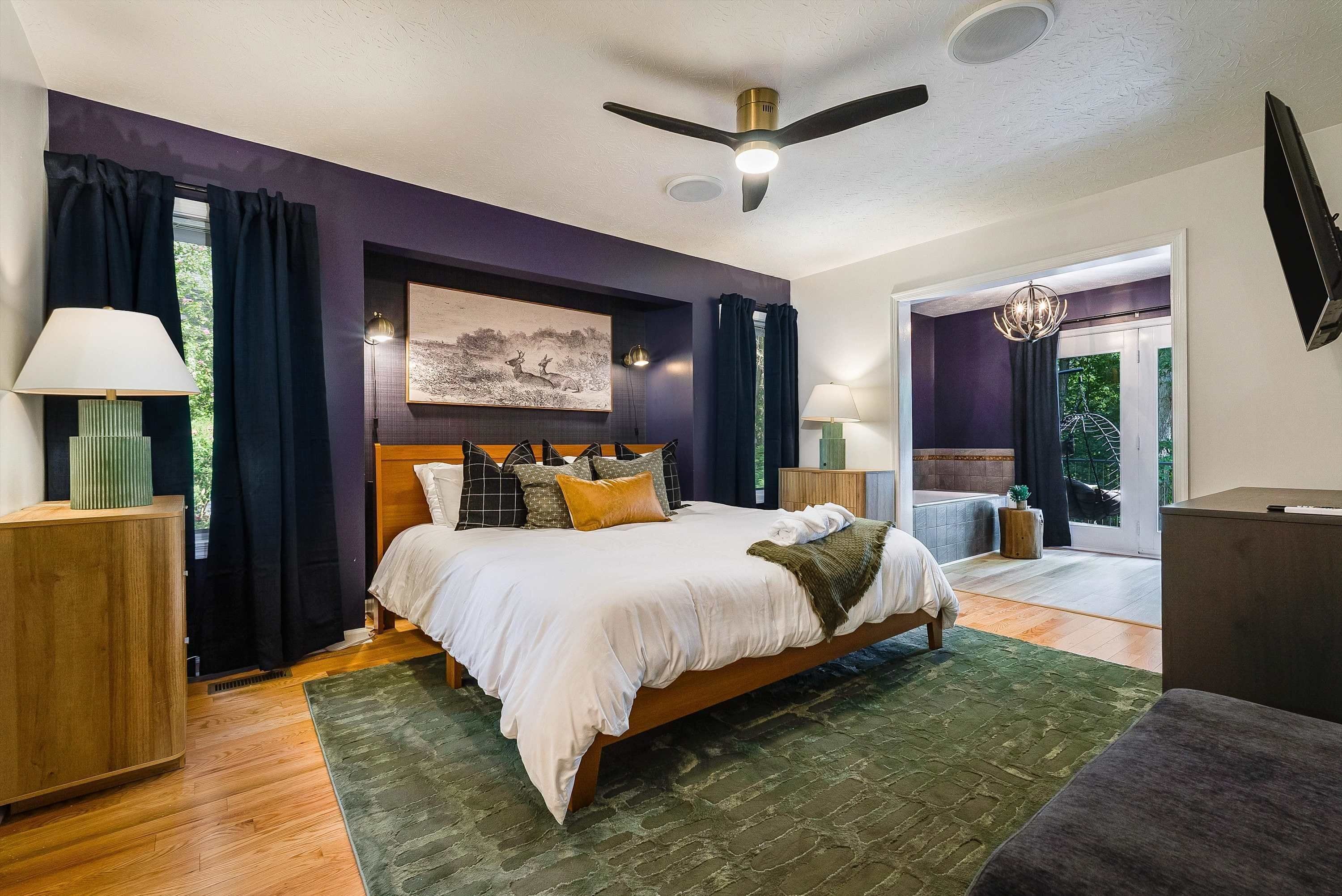 A bedroom with a bed, bedside lamps, a ceiling fan, a painting, and a view to the outside.