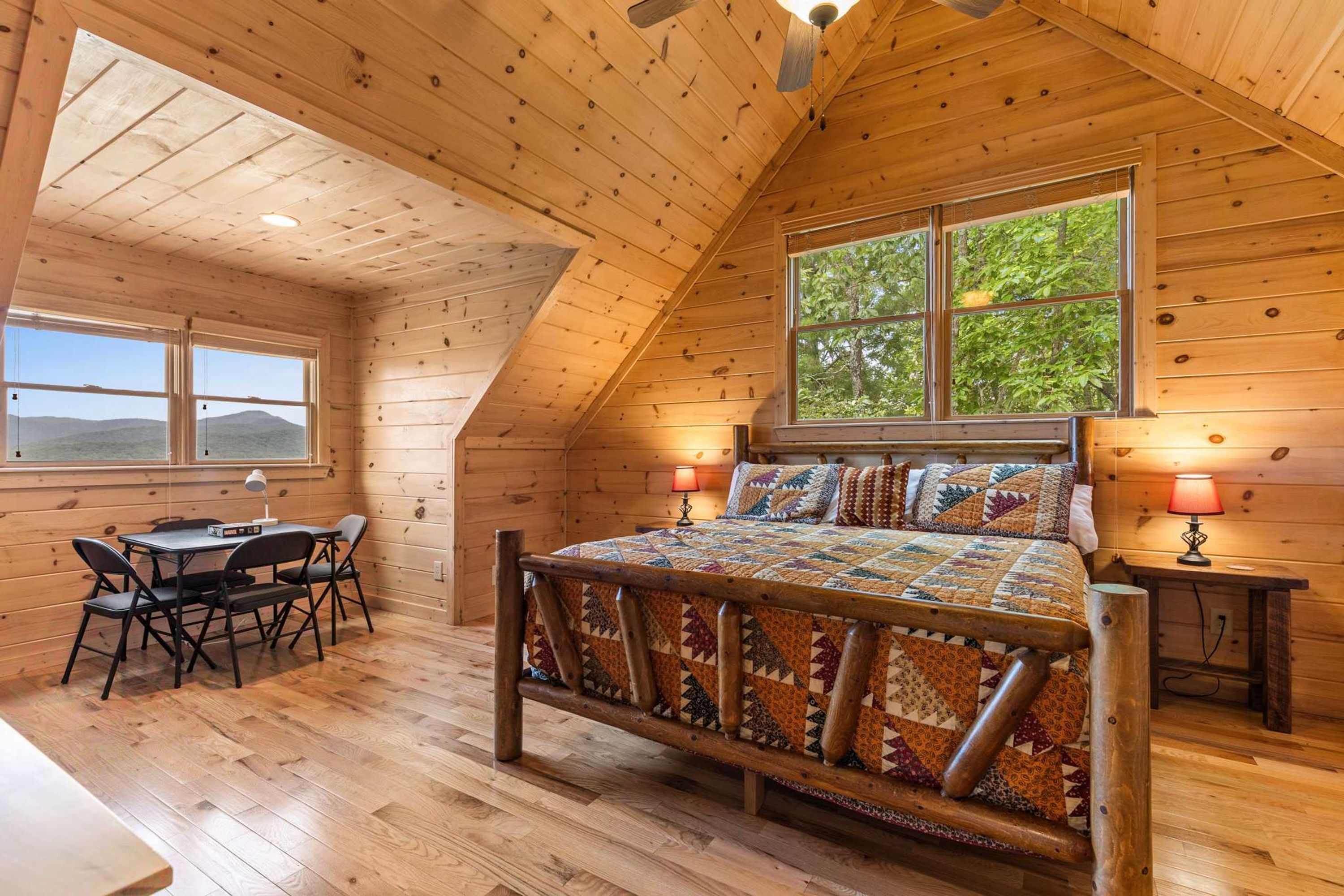 A wooden cabin with a loft bedroom, a dining area, and a large window offering a view of the outdoors.