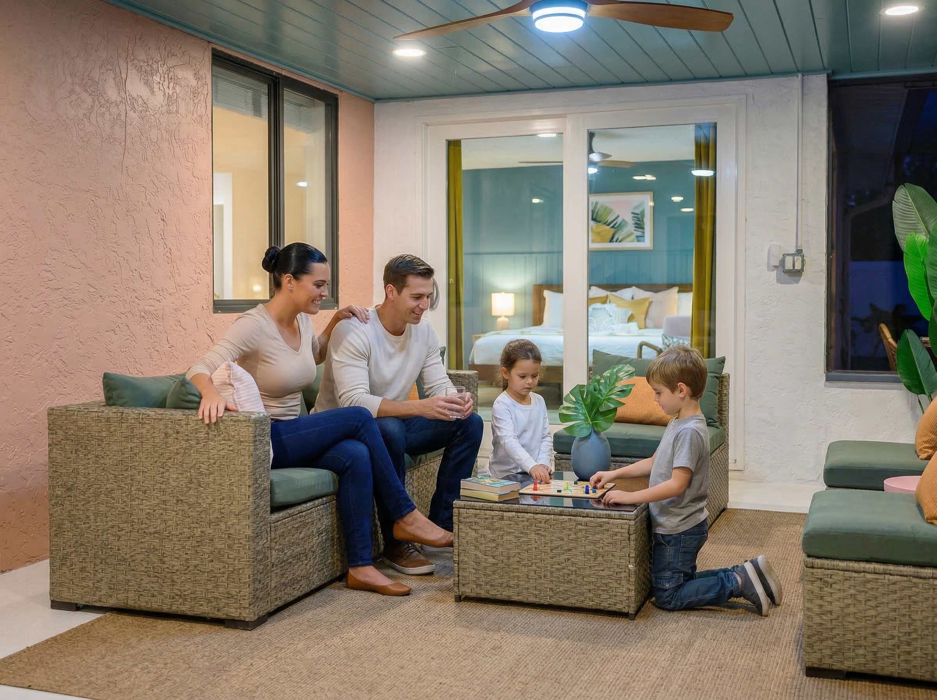 Una famiglia seduta su mobili in vimini in una stanza con una ventola a soffitto, una grande finestra e vista su una camera da letto.