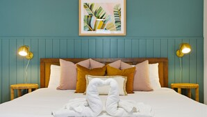 Ein ordentlich bezogenes Bett mit weißen Bettwäsche, eine Handtuch-Herz-Arrangement und ein gerahmtes Kunstwerk mit tropischen Blättern darüber.