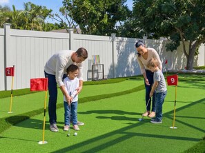 A family playing mini-golf on a green artificial turf course with numbered holes. - Poolside Oasis Sleeps 8 Game Room Putt Putt (Bradenton)