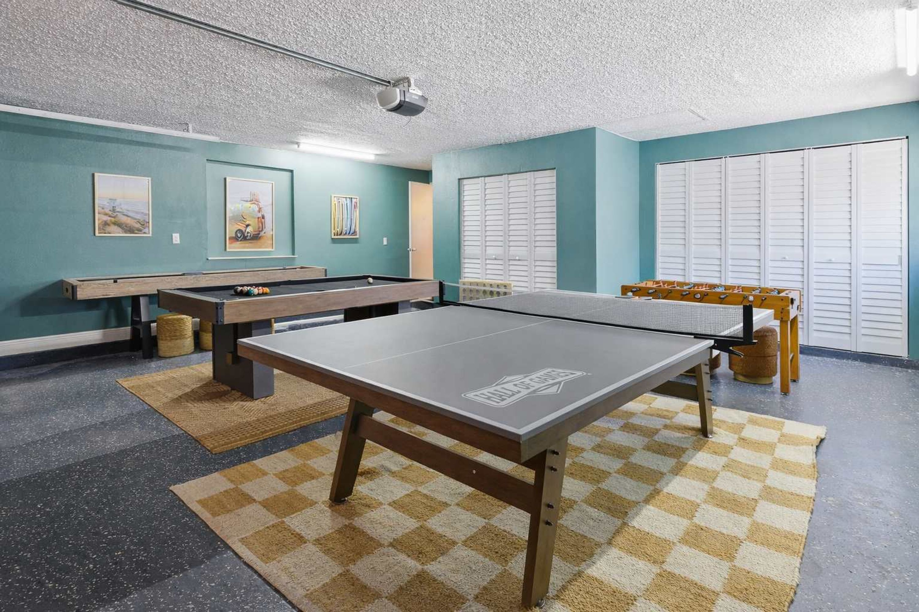 Een pooltafel in een kamer met turquoise muren, een aan het plafond gemonteerde projecteur en een geruit vloerkleed.