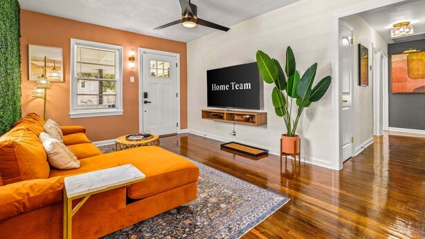 Una sala de estar con un sofá grande color naranja, un televisor de pantalla plana, un ventilador de techo y una planta de gran tamaño.
