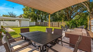 Un patio al aire libre con mesa y sillas, una zona de estar cubierta y vistas a un jardín con césped y árboles.