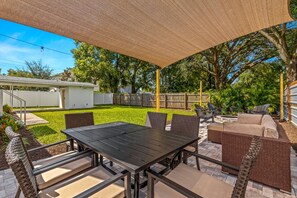 Ein Terrassenbereich im Freien mit Tisch und Stühlen, ein überdachter Sitzbereich und Blick auf einen Garten mit Rasen und Bäumen.