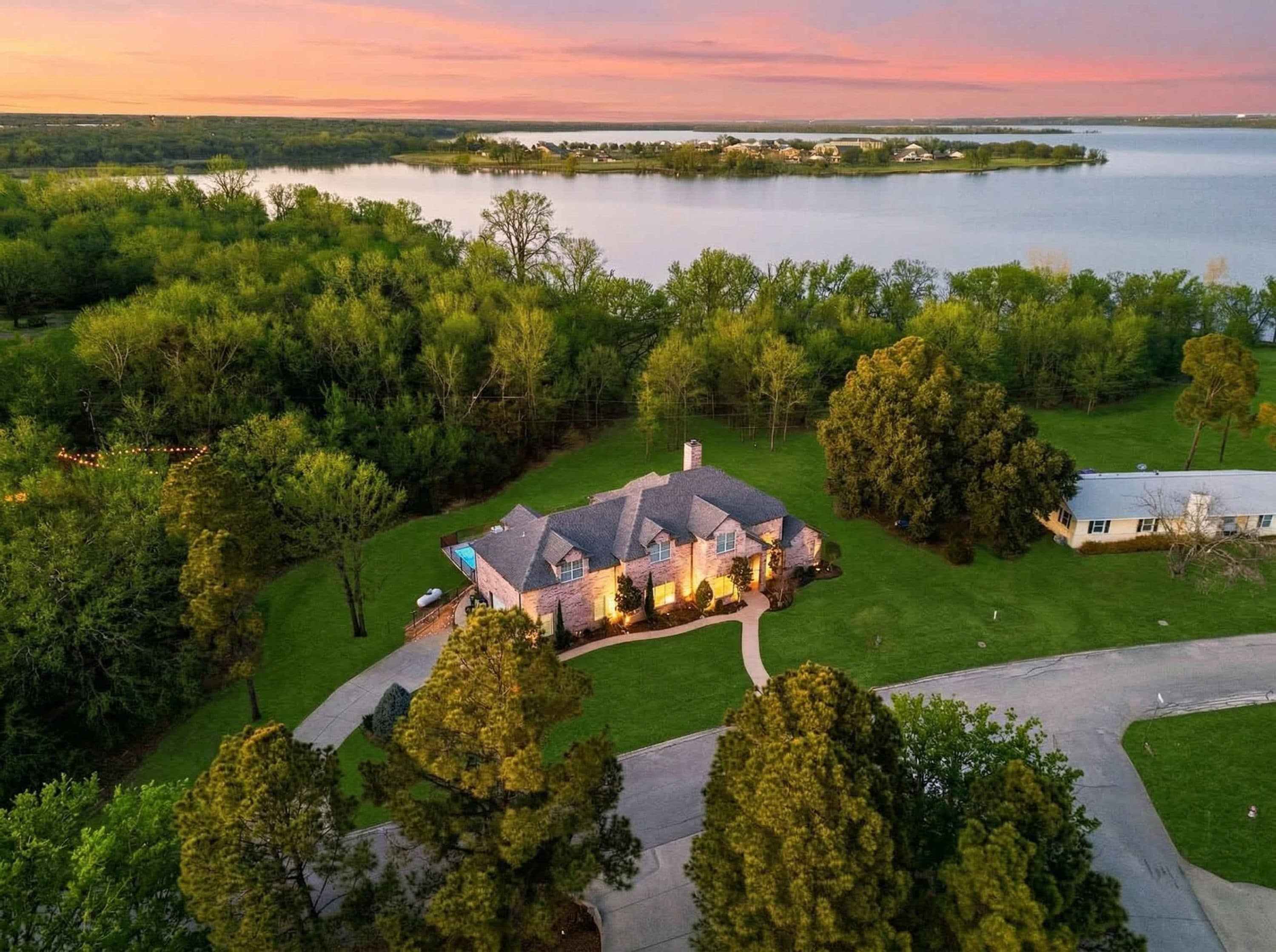 A large house with a stone exterior, surrounded by a well-maintained lawn and trees, with a lake in the background.