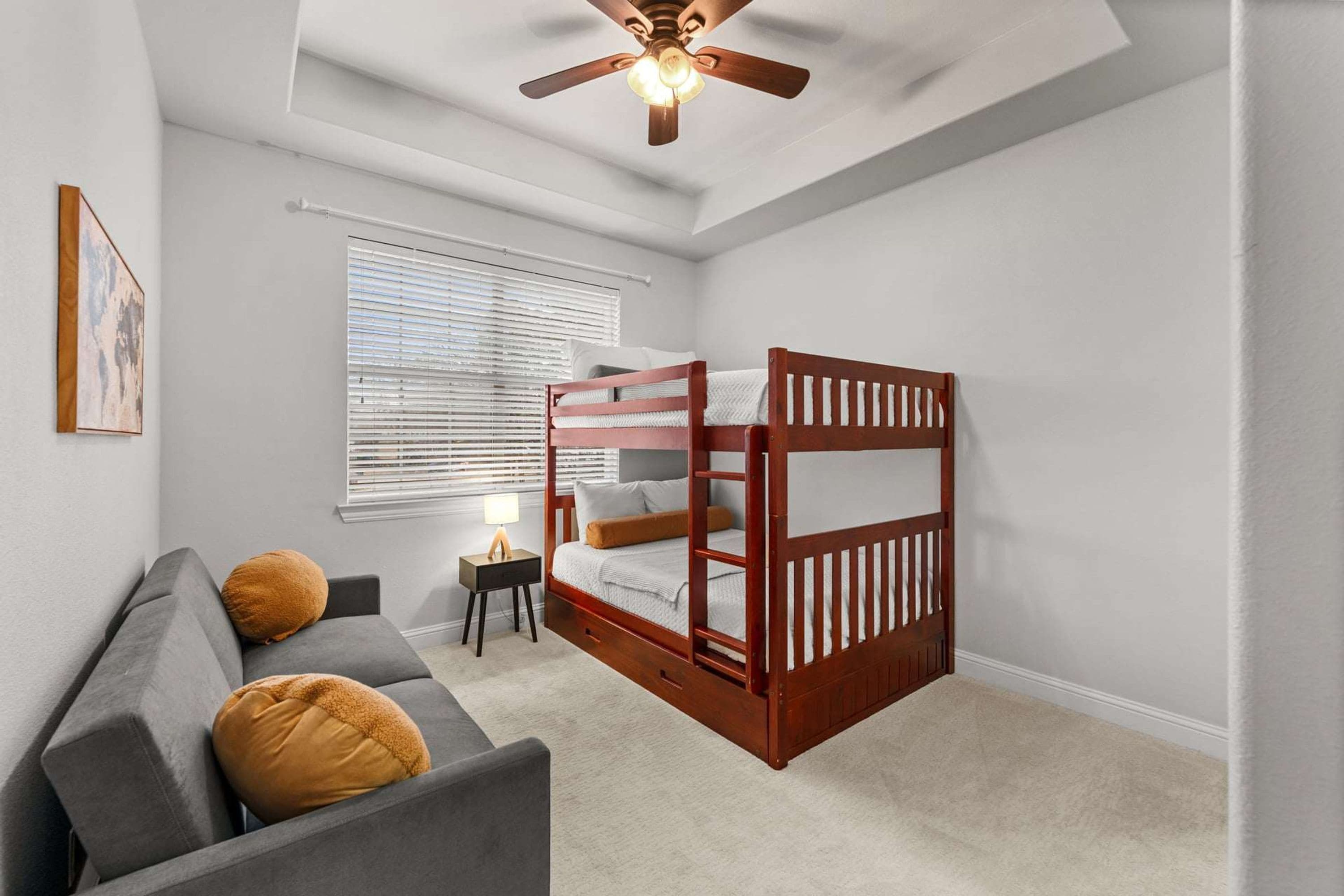 A bedroom with a bunk bed, a gray sofa, a small bedside table, and a ceiling fan.