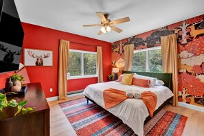 Une chambre avec un mur rouge, un lit avec une literie blanche, un ventilateur de plafond et une grande fenêtre avec des rideaux.