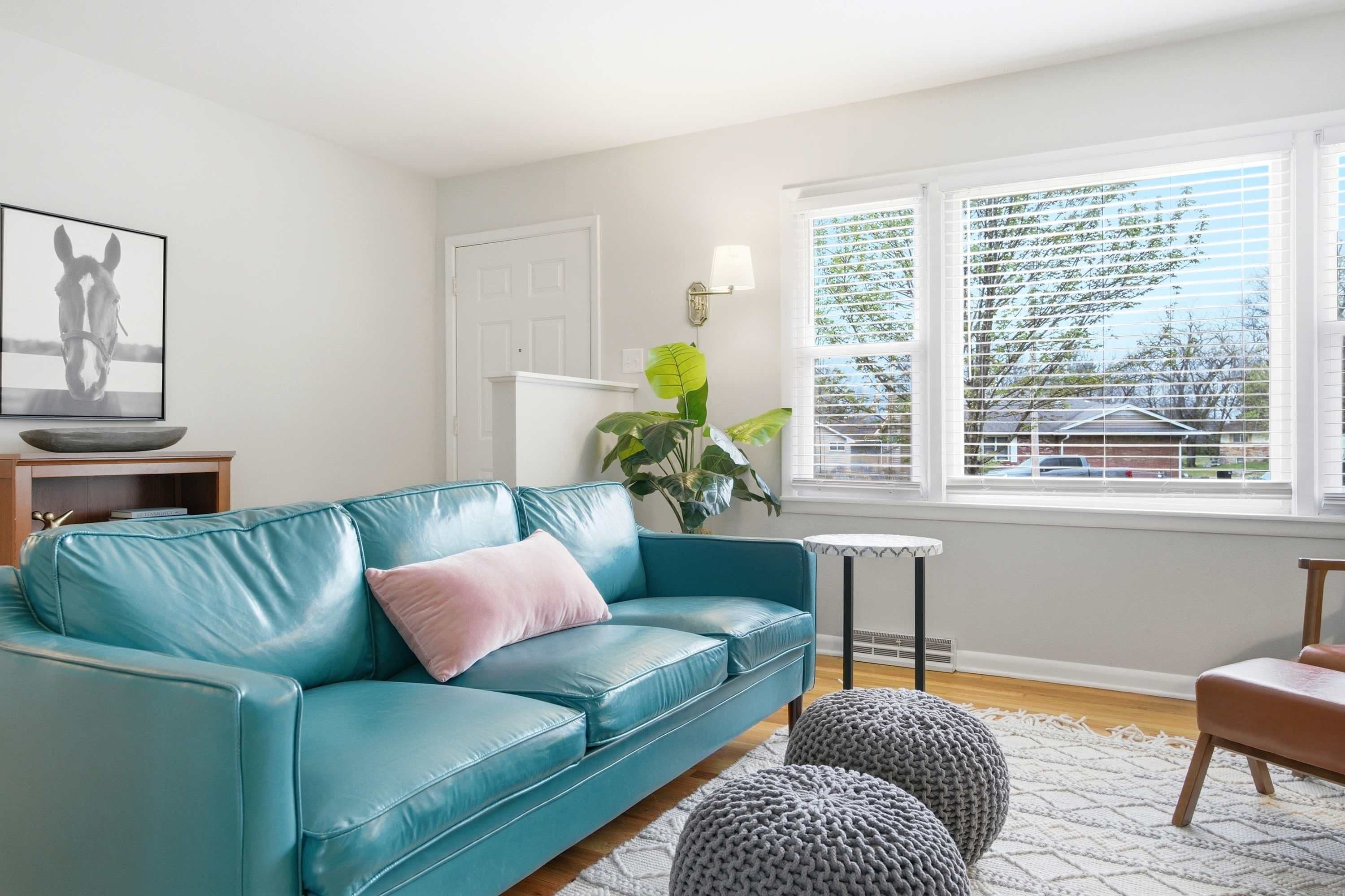 A living room with a teal leather sofa, a small round table, and a large window with blinds.