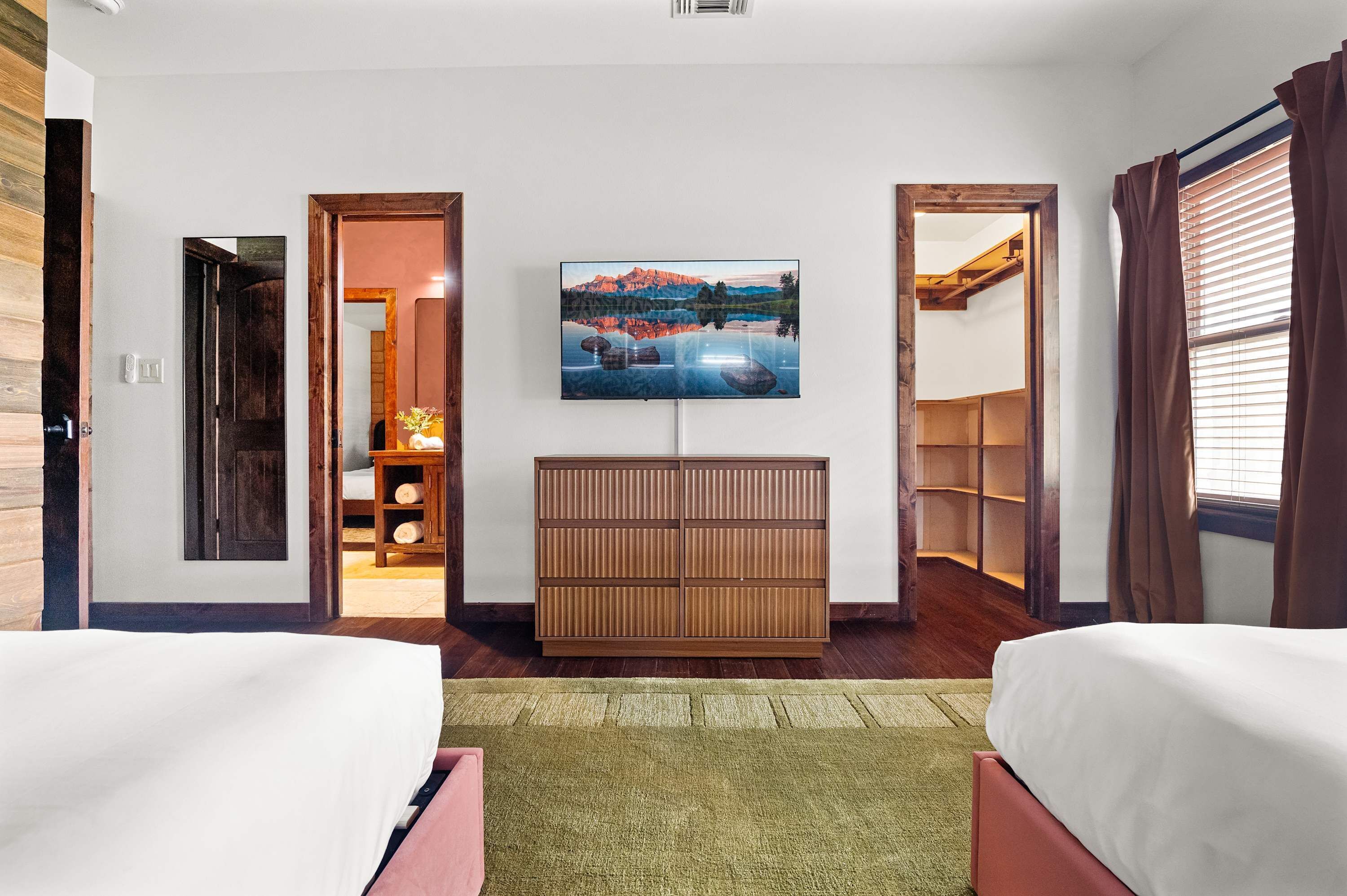 A hotel room with two beds, a TV on the wall, a wooden cabinet, and a view of a mountain landscape.