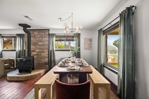 Un comedor con chimenea de piedra, una mesa de madera dispuesta para cuatro personas y amplios ventanales con vistas al exterior.