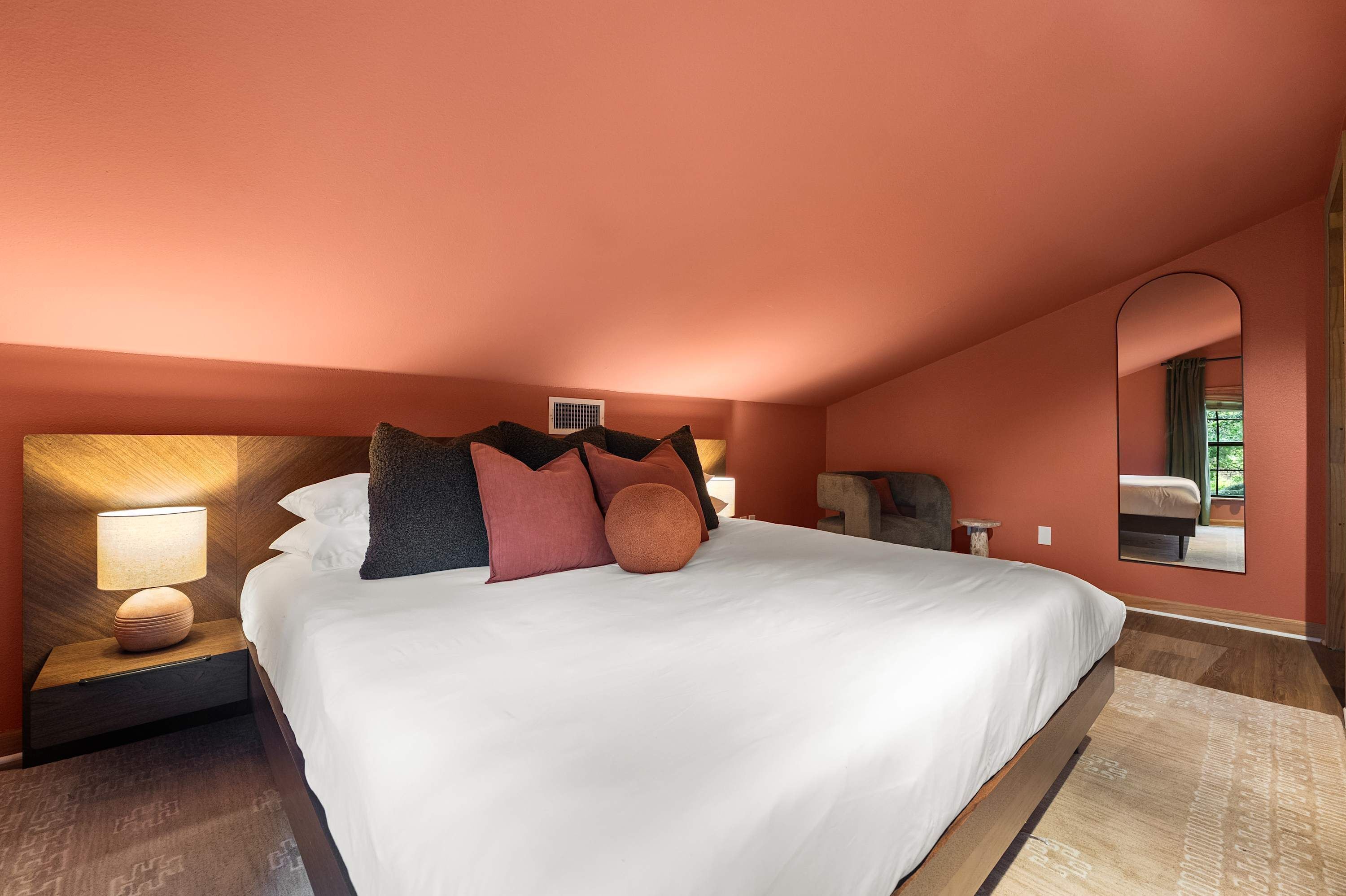 A hotel room with a large bed, a nightstand with a lamp, a mirror, and a window with a view of greenery.