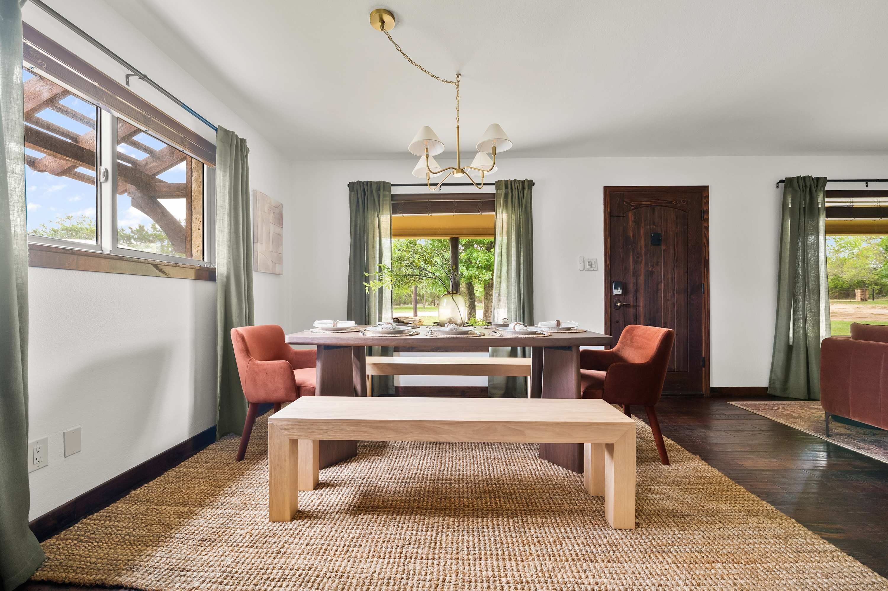 Un comedor con una mesa de madera, sillas y un banco. Hay una ventana con cortinas, una puerta y una lámpara de araña.