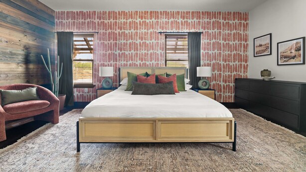 A bedroom with a large bed, two bedside tables, a wooden headboard, a red armchair, and a patterned wall. - Birdie Creek Ranch - Private Golf Course Resort (Driftwood)