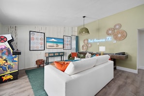 A living room with a white sofa, a gaming console, framed artwork, and a neon sign. - The Wave House Sleeps 16 Pool Hot Tub Bikes (Port Aransas)