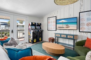 Ein Zimmer mit einer Couch, einem Couchtisch, Spielautomaten, einem Strandbild und Blick ins Freie.