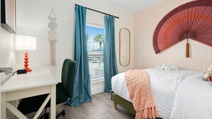 Una camera d'albergo con un letto, una scrivania, una sedia, una finestra con tende, un ventilatore a parete e un elemento decorativo.