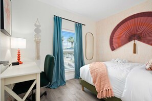 Ein Hotelzimmer mit Bett, Schreibtisch, Stuhl, einem Fenster mit Vorhängen, einem Wandventilator und einer Wanddekoration.