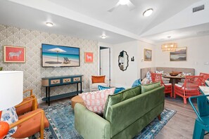Uma sala de estar moderna com um sofá verde, uma área de jantar com cadeiras vermelhas e um quadro com tema praiano na parede.