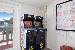Duas máquinas de fliperama com a marcação 'X-Men '97', uma parede branca com tinta descascada e uma obra de arte com tema de praia emoldurada.