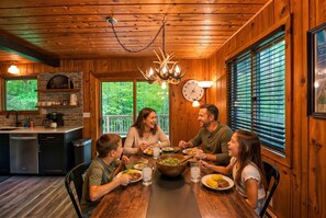 Une famille est réunie autour d’une table dans un chalet au décor intérieur en bois, avec une cheminée en briques et une vue sur la forêt par de grandes fenêtres.
