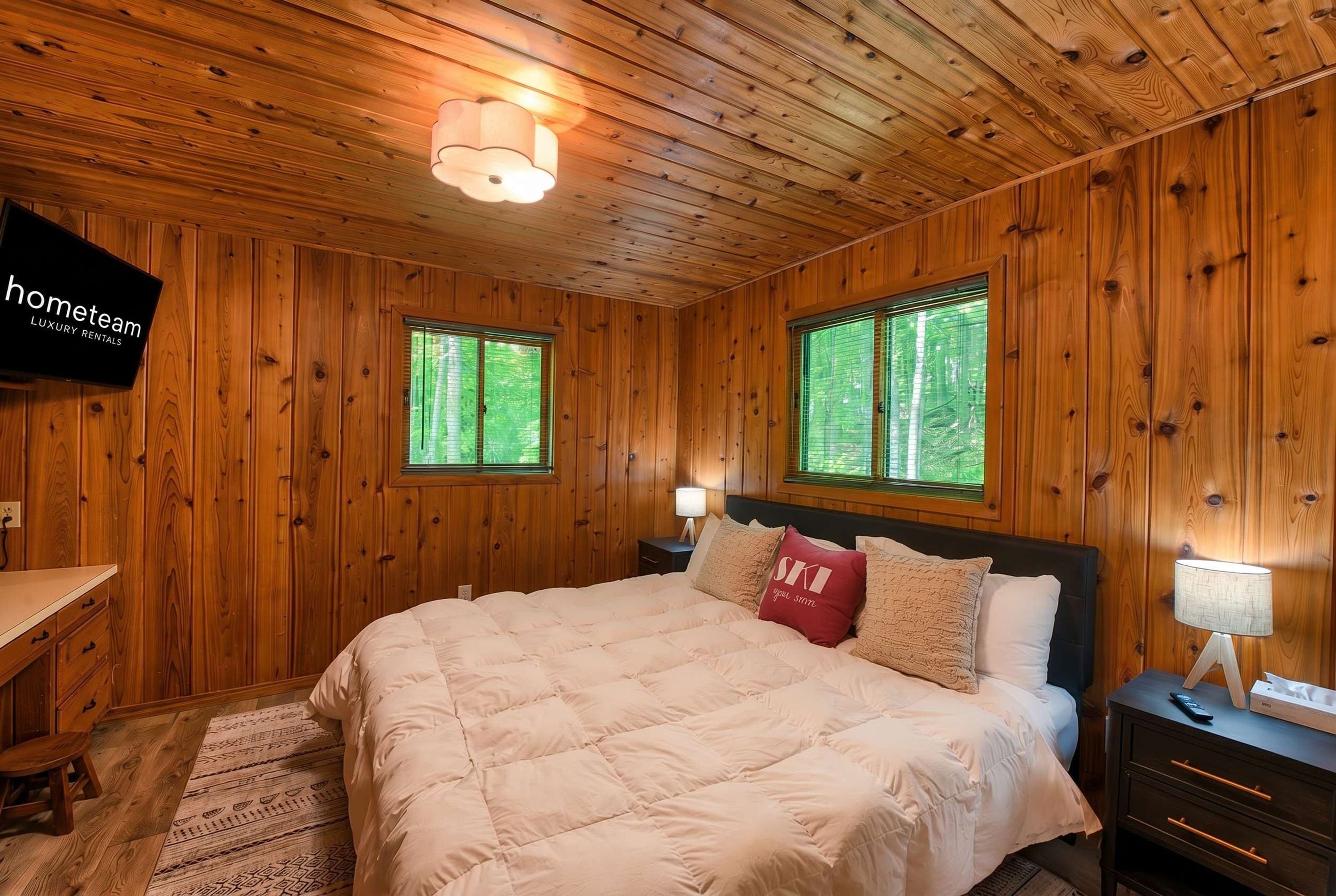 Dormitorio con paneles de madera, una cama, mesitas de noche, un escritorio y un televisor que muestra 'hometeam LUXURY RENTALS'.