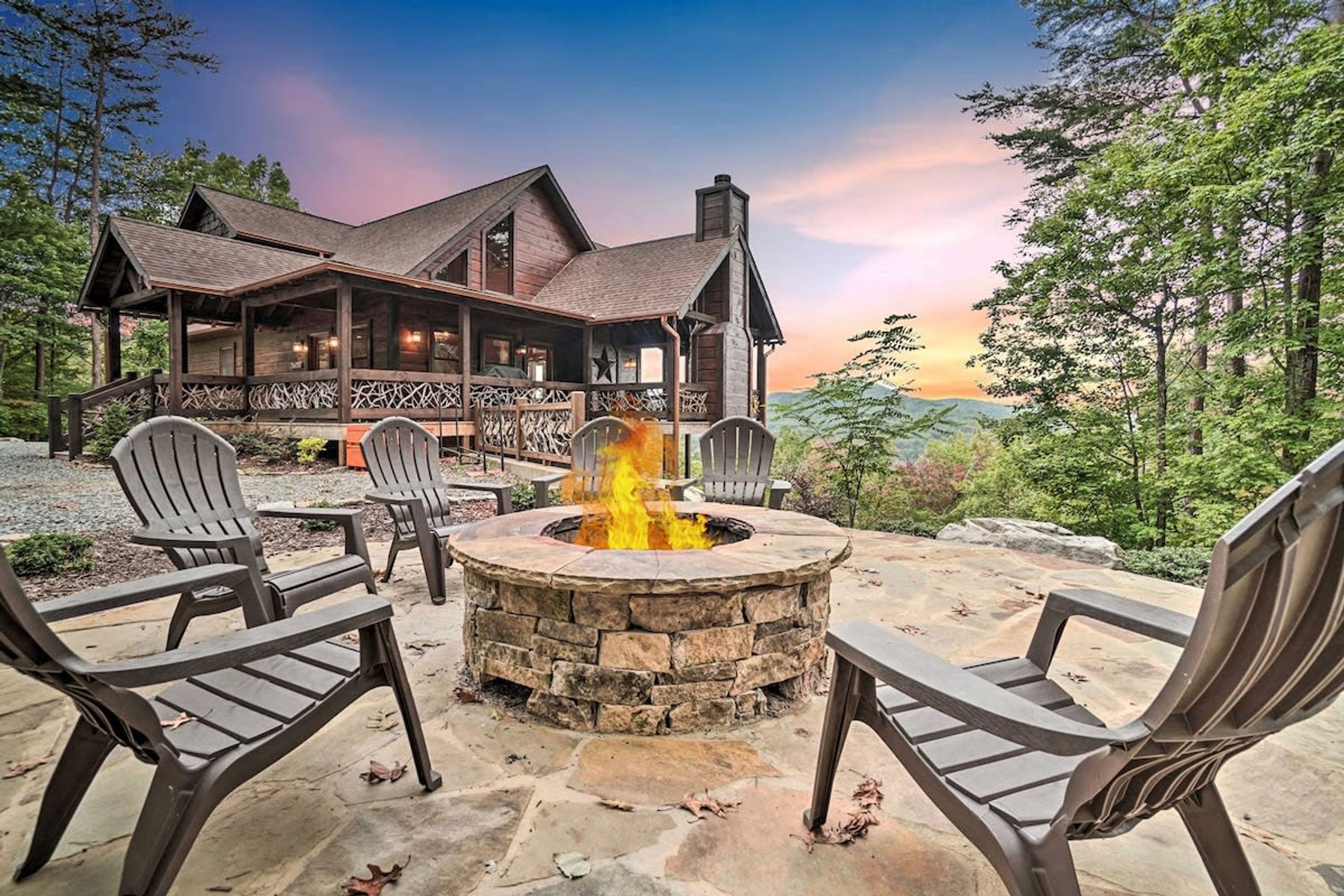 A lodge with a stone fire pit surrounded by Adirondack chairs, set against a backdrop of a large wooden house and a forested landscape.