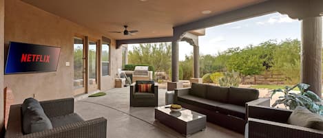 A covered outdoor living area with wicker furniture, a flat-screen TV, and a ceiling fan.