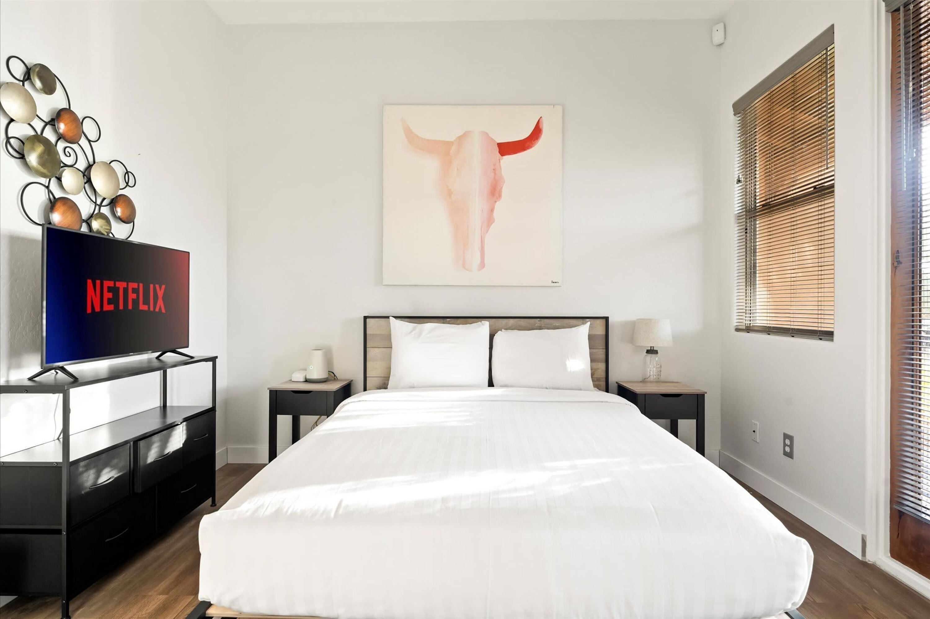 A modern hotel room with a large bed, a flat-screen TV, a decorative wall piece, and a window with blinds.